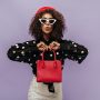 Trendy curly haired girl with white sunglasses and red cap in long sleeve blouse looking into camera and holding red handbag.