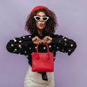 Trendy curly haired girl with white sunglasses and red cap in long sleeve blouse looking into camera and holding red handbag.