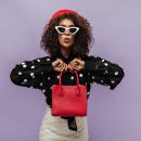 Trendy curly haired girl with white sunglasses and red cap in long sleeve blouse looking into camera and holding red handbag.